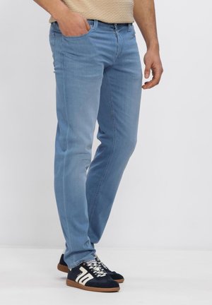 Mann trägt hellblaue Slim-Fit-Jeans und schwarz-weiße Turnschuhe, steht mit einer Hand in der Tasche vor weißem Hintergrund.