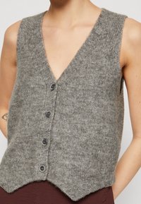 Szary bezrękawnik wykonany z miękkiego materiału, z głębokim dekoltem w kształcie V, pięcioma ciemnymi guzikami oraz wyprofilowanym dołem z teksturowanym wykończeniem.