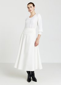 Weißer, langer Rock mit tailliertem Bund und strukturiertem Strickoberteil. Glattes Material, fließendes Design und Dreiviertelärmel. Schwarze spitze Stiefel ergänzen den Look.