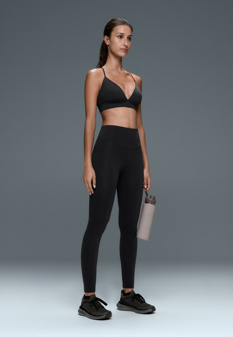 Frau in schwarzem Sport-BH und Leggings hält eine Wasserflasche, steht vor einem grauen Hintergrund und trägt schwarze Sportschuhe.