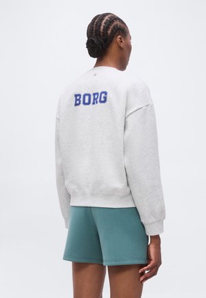 Person with braided hair wearing a light grey sweatshirt with "BORG" text on the back and teal shorts, standing against a plain background.
