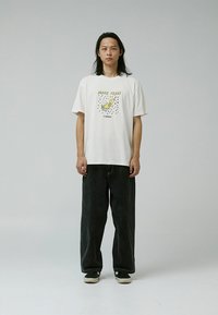 Tricou alb din bumbac cu un grafic de pasăre galbenă, asortat cu pantaloni negri din denim largi. O pereche de încălțări negre simple completează ținuta.