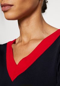 Granatowy sweter z głębokim dekoltem w serek, z kontrastującym czerwonym ściągaczem. Gładki materiał o dopasowanym kroju.