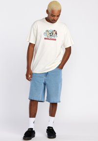 Camiseta de algodón color crema con un estampado gráfico que presenta personajes y texto, combinada con pantalones cortos de mezclilla azul claro y zapatillas negras.