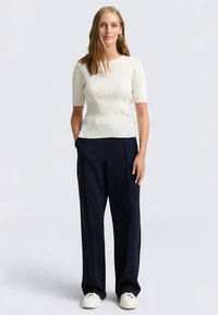 Roomkleurige geribbelde gebreide top met korte mouwen en een nauwsluitend silhouet, gecombineerd met marineblauwe broek met wijde pijpen en witte sneakers.