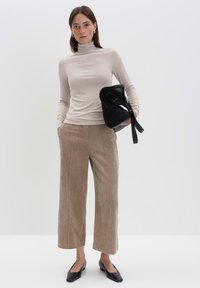 Helle beige Rollkragenbluse kombiniert mit taubenfarbigen, weiten Cordhosen. Das Model hält eine schwarze strukturierte Tasche und trägt schwarze spitze Ballerinas.