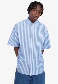 Camicia a maniche corte con bottoni di colore azzurro chiaro, con righe verticali bianche e rosa. Presenta un colletto e un'etichetta sul petto a sinistra.