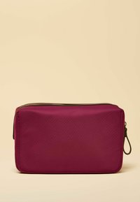 Prostokątna fioletowa kosmetyczka o gładkiej, nylonowej fakturze, z zamkiem błyskawicznym na górze i małą boczną pętlą ułatwiającą trzymanie.
