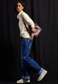 Frau, die mit der Hand in der Tasche läuft, trägt hochgekrempelte Jeans, einen weißen Rollkragenpullover, weiße Sneakers, eine burgunderrote Ledertasche und ein passendes Armband.