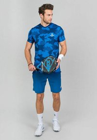 Camiseta atlética de camuflaje azul, pantalones cortos azules, muñequera blanca, calcetines blancos y zapatillas deportivas blancas; sosteniendo una pala de pádel negra y azul.