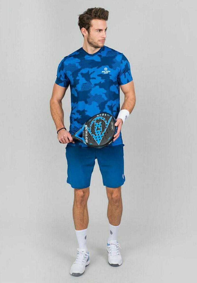 Camiseta atlética de camuflaje azul, pantalones cortos azules, muñequera blanca, calcetines blancos y zapatillas deportivas blancas; sosteniendo una pala de pádel negra y azul.