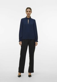Marineblauwe blouse met lange mouwen, een gerimpelde kraag en een keyhole voorkant, gecombineerd met zwarte wijde broekspijpen en puntige hakken.