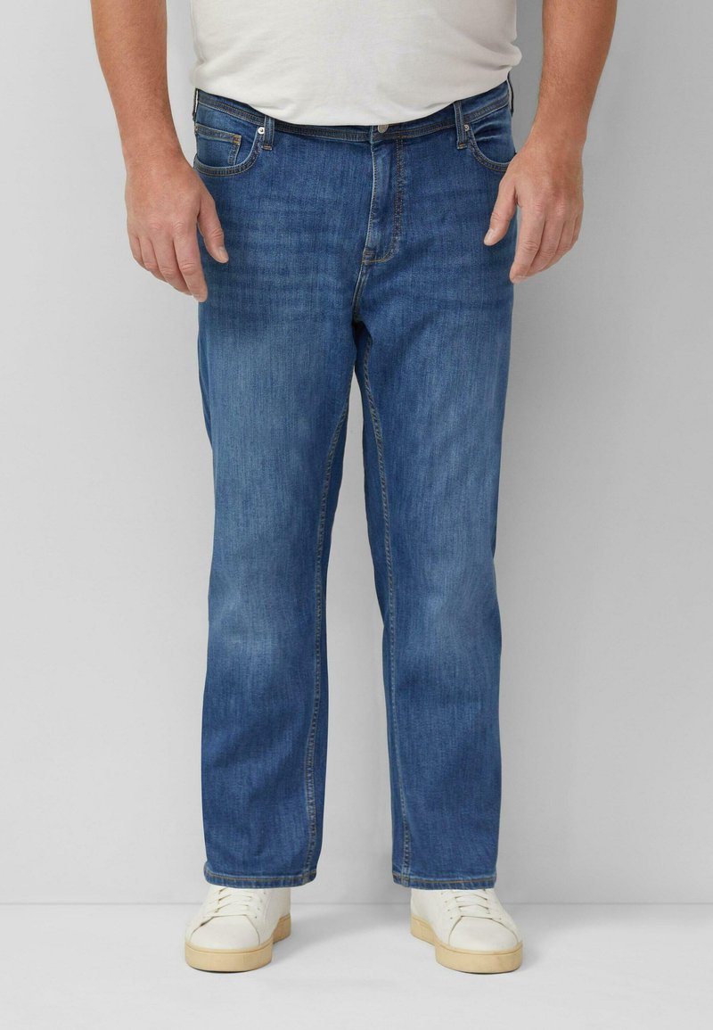 Mann trägt blaue Jeans mit geradem Bein, weiße Turnschuhe und ein weißes, in die Hose gestecktes Hemd, steht vor einem einfarbigen grauen Hintergrund.