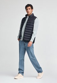 Czarna kamizelka puchowa z poziomymi przeszyciami, noszona na jasnoszarym swetrze, w zestawie z jasnoniebieskimi dżinsami i beżowymi trampkami.