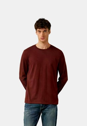 Junger Mann mit kurzen lockigen Haaren, der ein schlichtes dunkelrotes Langarmshirt und blaue Jeans trägt, vor einem hellgrauen Hintergrund stehend.