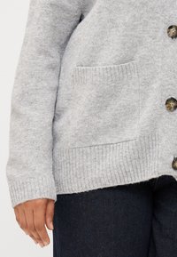 Gray cardigan with ribbed hems, featuring a single pocket and buttons, worn over dark denim pants, highlighting a hand resting on the fabric.
