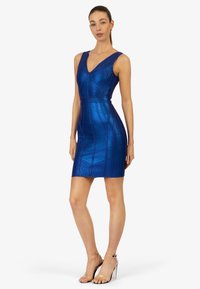 Robe sans manches bleue avec un col en V, fabriquée à partir d'un tissu texturé et brillant. Elle présente un design ajusté et des motifs géométriques, accompagnée de talons transparents.