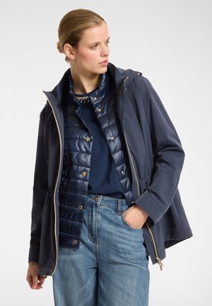 Giacca impermeabile blu navy con cappuccio sopra un gilet imbottito blu navy, con dettagli in metallo dorato e chiusura con zip. Indossata con jeans in denim azzurro.