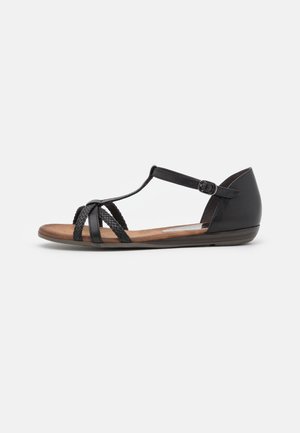 Sort flad sandal med ankelsrem, flettede frontremme og tan polstret sål på hvid baggrund.