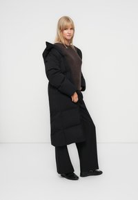 Manteau rembourré noir au design long et surdimensionné, fabriqué en matériau brillant. Doté d'une capuche, d'un motif matelassé et de poignets côtelés.