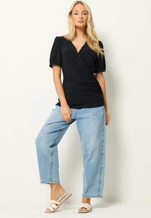 Kvinde stående foran en ensfarvet baggrund iført en sort slå-om-bluse, lyseblå wide-leg jeans og hvide slippers.