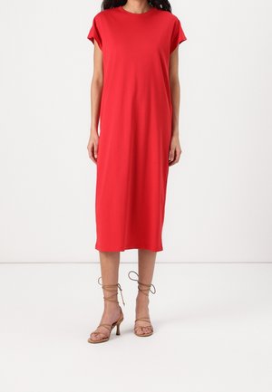 Mujer vestida con un vestido rojo liso de manga corta y largo midi, con sandalias de tacón con tiras en color marrón claro, posando frente a un fondo blanco liso.