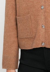 Cardigan in lana marrone chiaro con polsini e orlo a coste, dotato di una tasca e dettagli con bottoni argentati. Pantaloni neri parzialmente visibili.