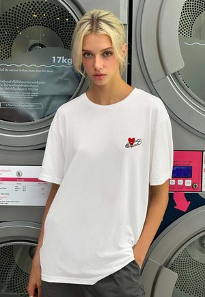 T-shirt en coton blanc avec un design de cœur rouge et d'épingle de sûreté sur la poitrine, doté d'une coupe décontractée et de manches courtes.