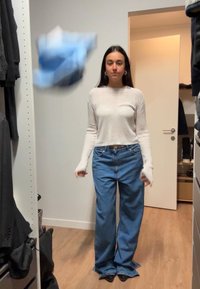 Witte geribde trui met lange mouwen, blauwe wijde jeans en zwarte hakken; houten vloer en kast op de achtergrond.