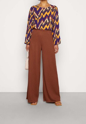 Long-sleeve blouse with a purple, yellow, and cream zigzag pattern, paired with wide-leg brown trousers and tan sandals.