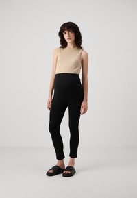 Μπεζ ριμπ αμάνικο τοπ συνδυασμένο με μαύρες, ελαστικές κολάν με ψηλή μέση. Το μοντέλο φορά μαύρες παντόφλες. Απλό φόντο.