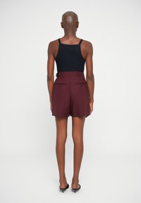 Bordowe spersonalizowane szorty z tylnymi kieszeniami, noszone z czarnym topem bez rękawów. Gładki materiał, długość do połowy uda i minimalistyczne detale projektowe.