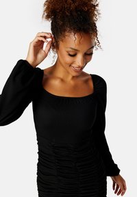 Czarna bluzka z długim rękawem z kwadratowym dekoltem, o żebrowanej fakturze i marszczeniami po bokach, uzupełniona o ozdobne kolczyki.