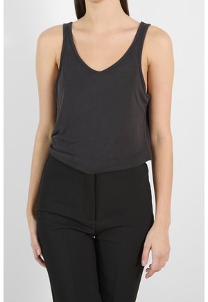 Kvinde iført en ærmeløs mørkegrå V-hals top og sorte bukser, stående mod en ensfarvet lys baggrund.