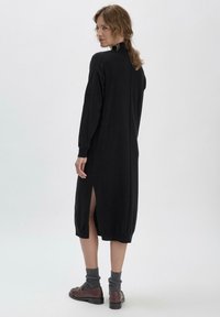 Schwarzes gestricktes Kleid mit langen Ärmeln, seitlichen Schlitzen und gerippten Bündchen. Der Stoff hat eine glatte Textur und wird mit grauen Socken und braunen Schuhen getragen.
