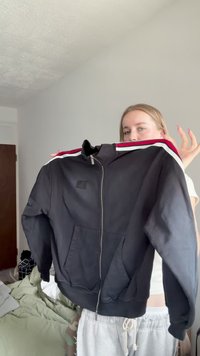 Personne tenant une veste noire zippée avec des bandes blanches et rouges sur les épaules dans une chambre.