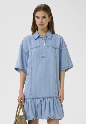Jeune femme portant une robe ample en jean bleu clair à manches courtes et ourlet volanté, tenant un sac à main marron sur un fond uni.