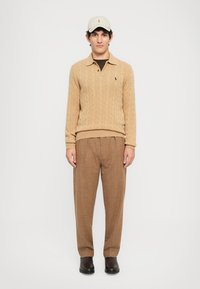 Pull en tricot beige avec un col, pantalon écossais marron, et une casquette de couleur claire, complétant un look coordonné.