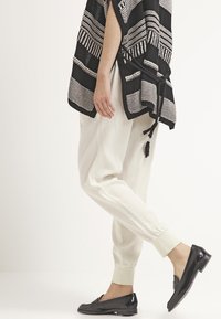 Poncho à motifs noir et blanc, coupe ample, associé à un pantalon crème fuselé et des mocassins noirs. Le tissu semble léger et texturé.