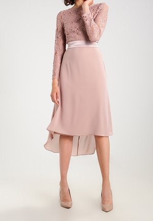 Femme portant un haut à manches longues en dentelle rose poudré associé à une jupe midi asymétrique et des chaussures à talons hauts couleur nude.