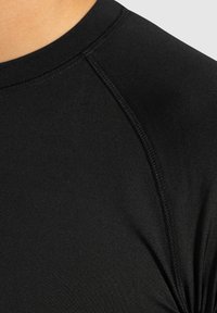 Schwarzes Sportshirt aus glattem, dehnbarem Material. Hat einen Rundhalsausschnitt und Raglanärmel mit Flachnahtsäumen sowie eine figurbetonte Silhouette.