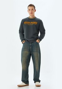 Sudadera gris oscuro con puños y cuello a rayas amarillas, que presenta texto gráfico en color naranja. Jeans de corte holgado, descoloridos, en azul y zapatillas blancas.