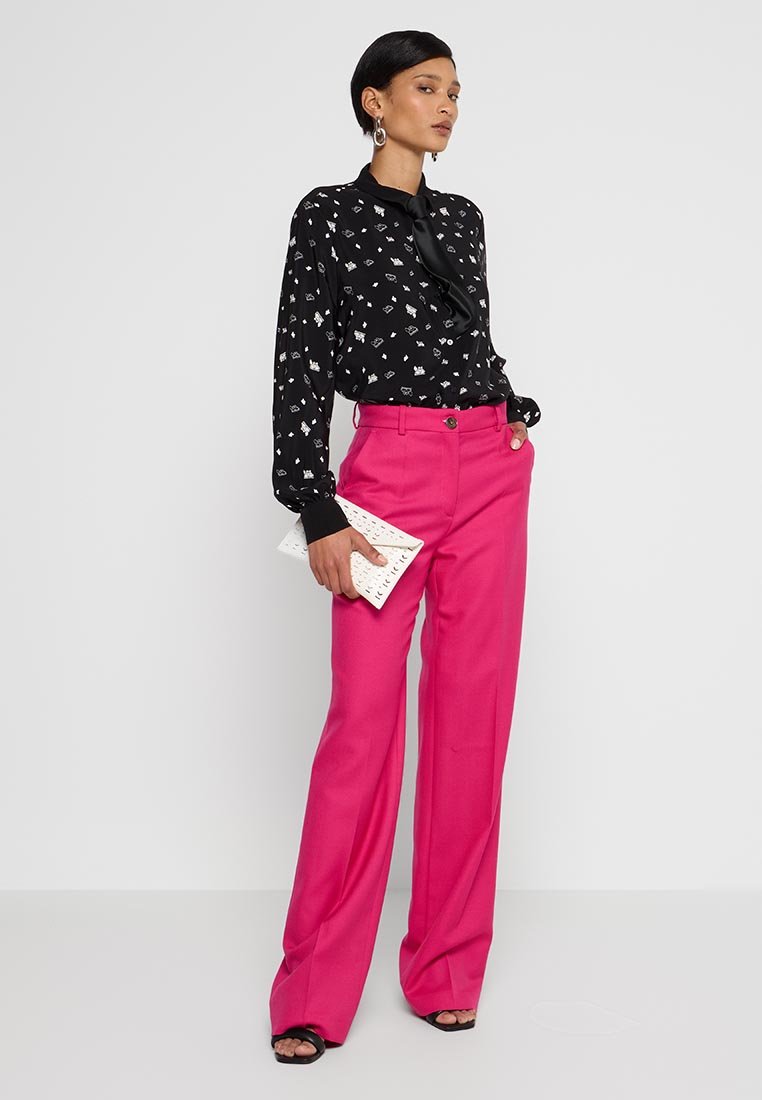 Camisa negra con patrones blancos y cuello de lazo, combinada con pantalones de pierna ancha de color rosa brillante. Sosteniendo un clutch blanco y usando tacones negros.