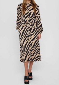 Beige und schwarze Kleid mit Zebramuster, Knopfleiste, langen Ärmeln, einem Gürteldetail in der Taille und in Midi-Länge. Getragen mit schwarzen Plattform-Sandalen.