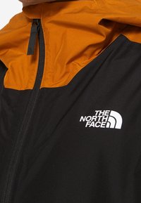 Czarna i pomarańczowa kurtka z przednim zamkiem błyskawicznym oraz białym logo The North Face na czarnej tkaninie w okolicy klatki piersiowej.