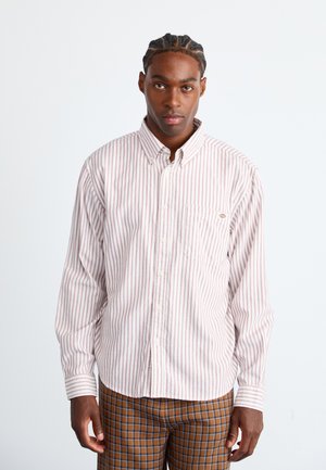 Man with braided hair wearing white and red striped button-up shirt and brown checkered pants, standing against white background.