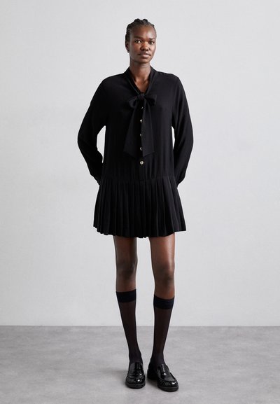 Robe noire avec une jupe plissée, nœud papillon au col, fermeture par boutons, manches longues. Associée à des chaussettes montantes et des mocassins.