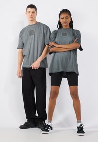 Graue, übergroße T-Shirts mit einem zentralen Grafikdesign, kombiniert mit schwarzen Hosen und Radhosen. Die Sneakers haben schwarze und weiße Designelemente.