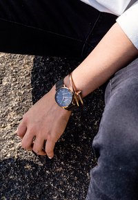 Reloj con correa de malla negra, esfera azul y detalles dorados, llevado en una mano. Diseño sencillo, caja redonda y silueta elegante.