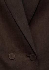 Blazer in lino marrone con rever a rotondo, bottoni doppi e superficie testurizzata. I bottoni sono circolari con una finitura liscia.